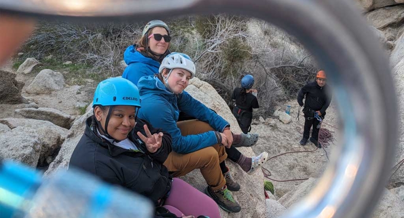 The photo is taken through the center of a carabiner, framing people wearing helmets sitting on a rock and smiling for the photo.    